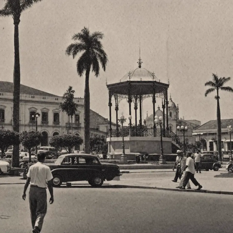 Praça da cidade nos anos 1950 com coreto e palmeiras, em preto e branco sépia